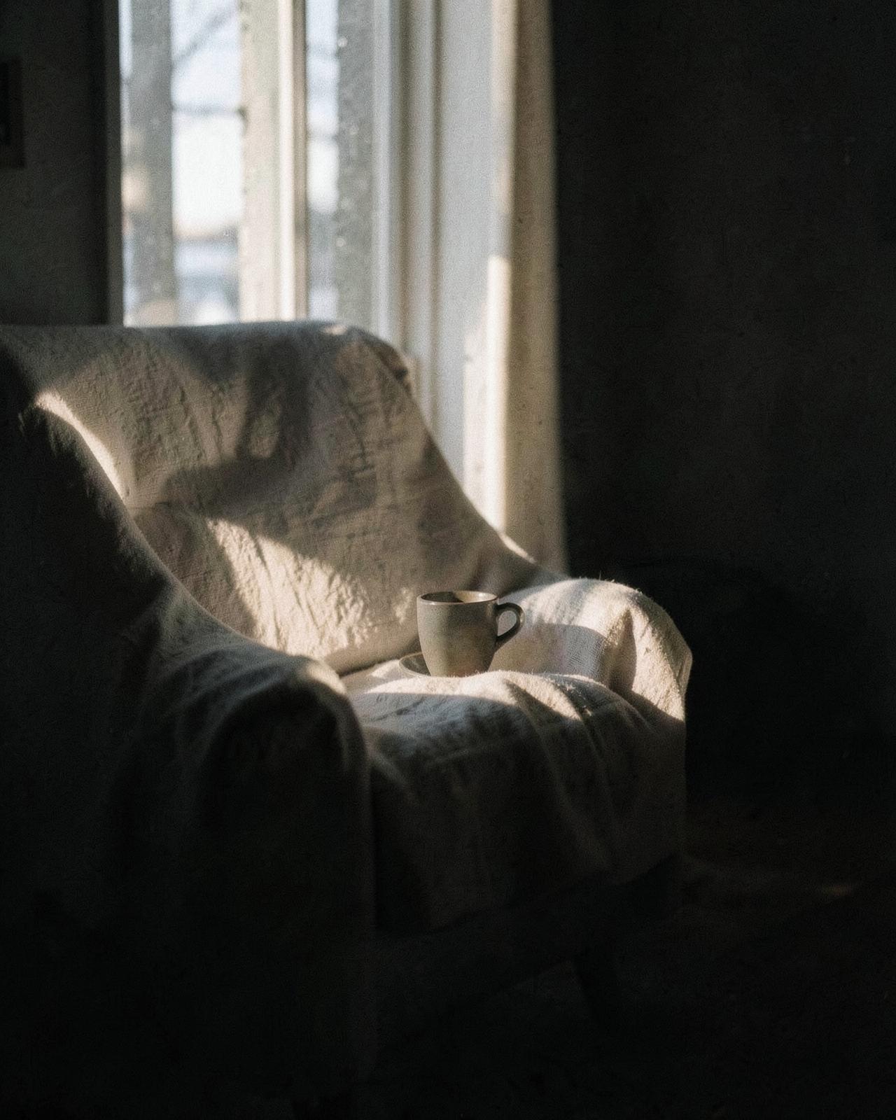 Winter morning light falling across a linen chair and a cup of coffee in a quiet dark room