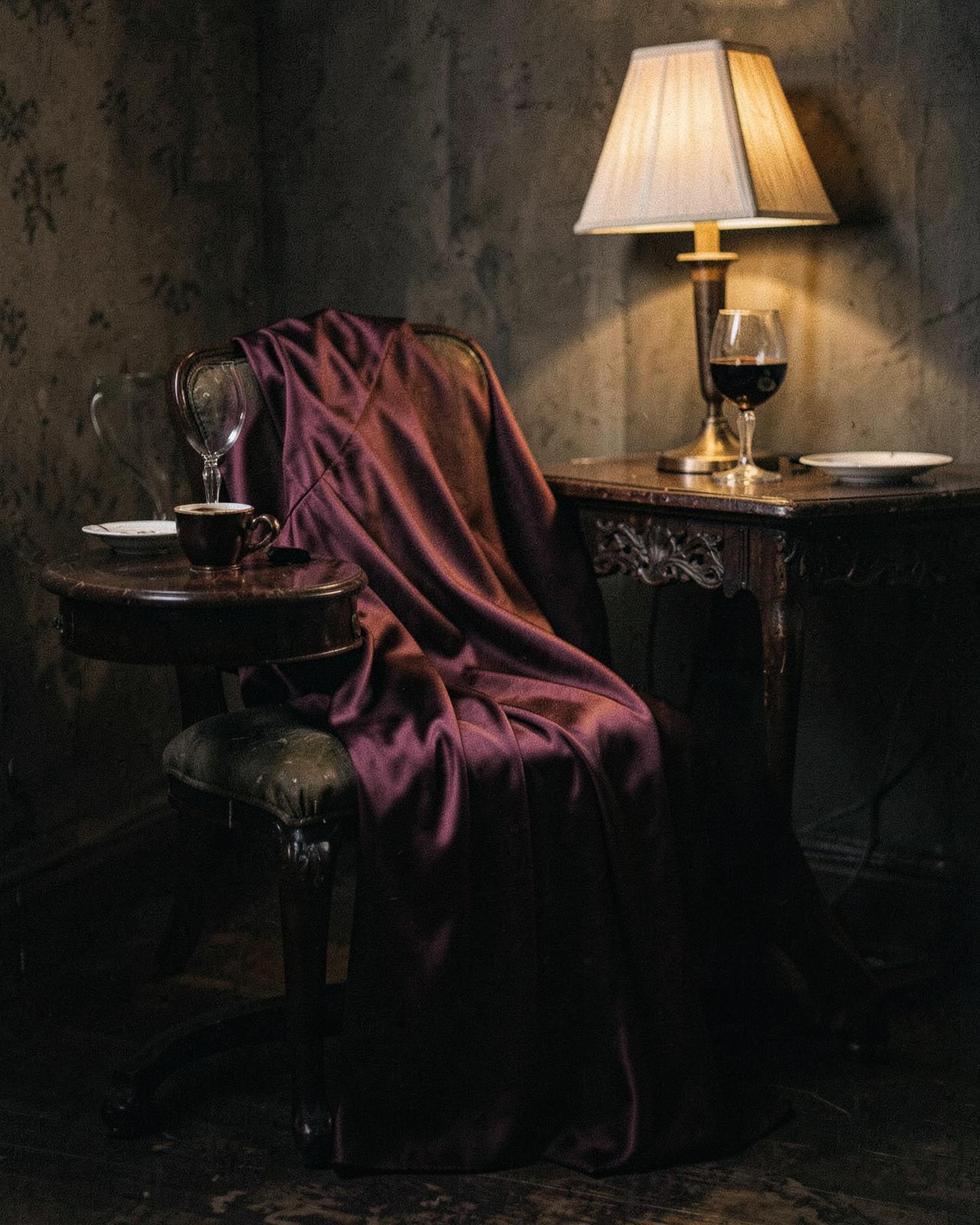 A burgundy silk dress draped over an antique chair beside a glass of wine in lamplight