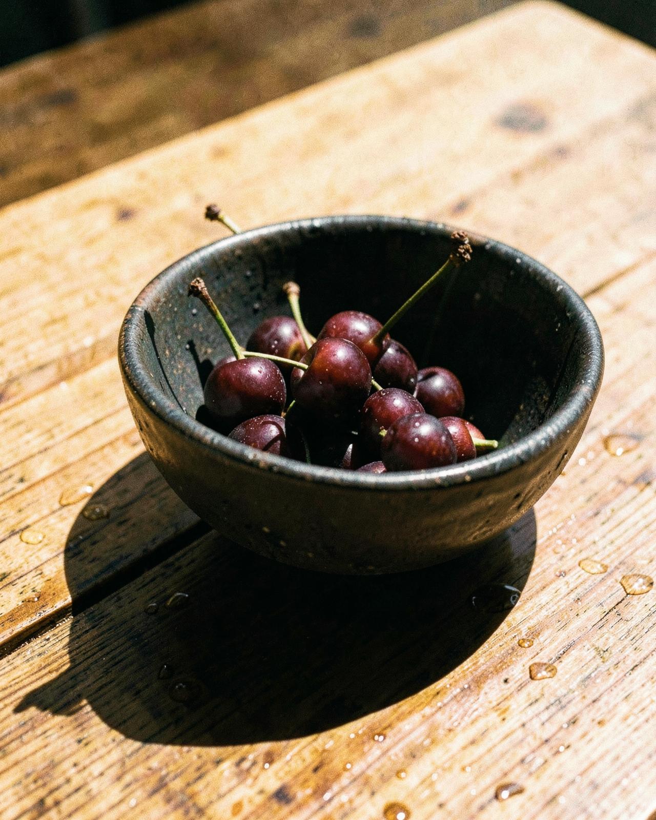 A dark bowl of cold cherries on a sunlit wooden table