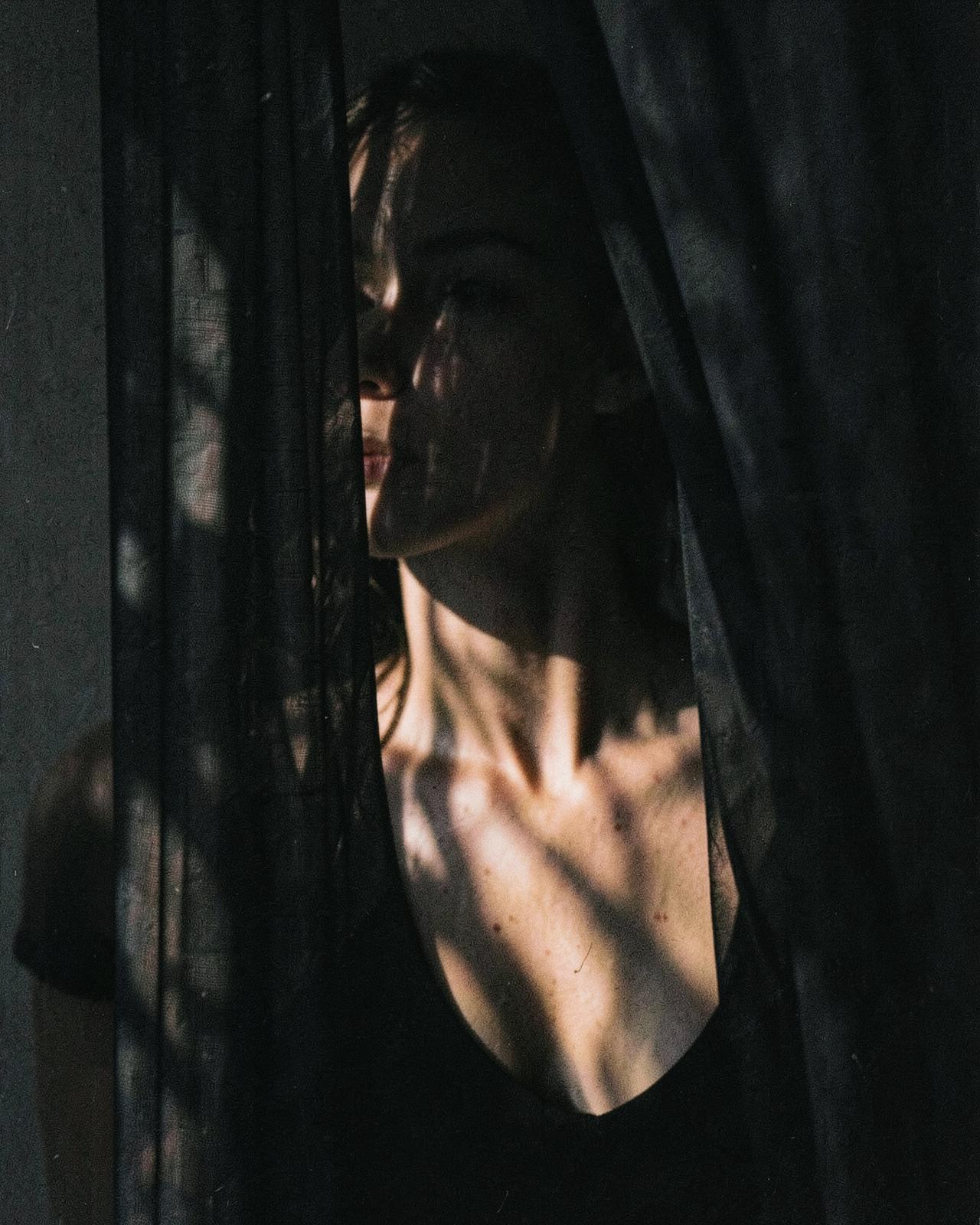 A woman half hidden behind a dark curtain, only her collarbone catching the light
